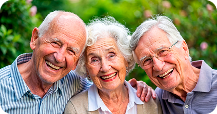 Three joyful elderly friends, two men and a woman, smiling broadly in a close embrace outdoors, depicting happiness and companionship in their later years